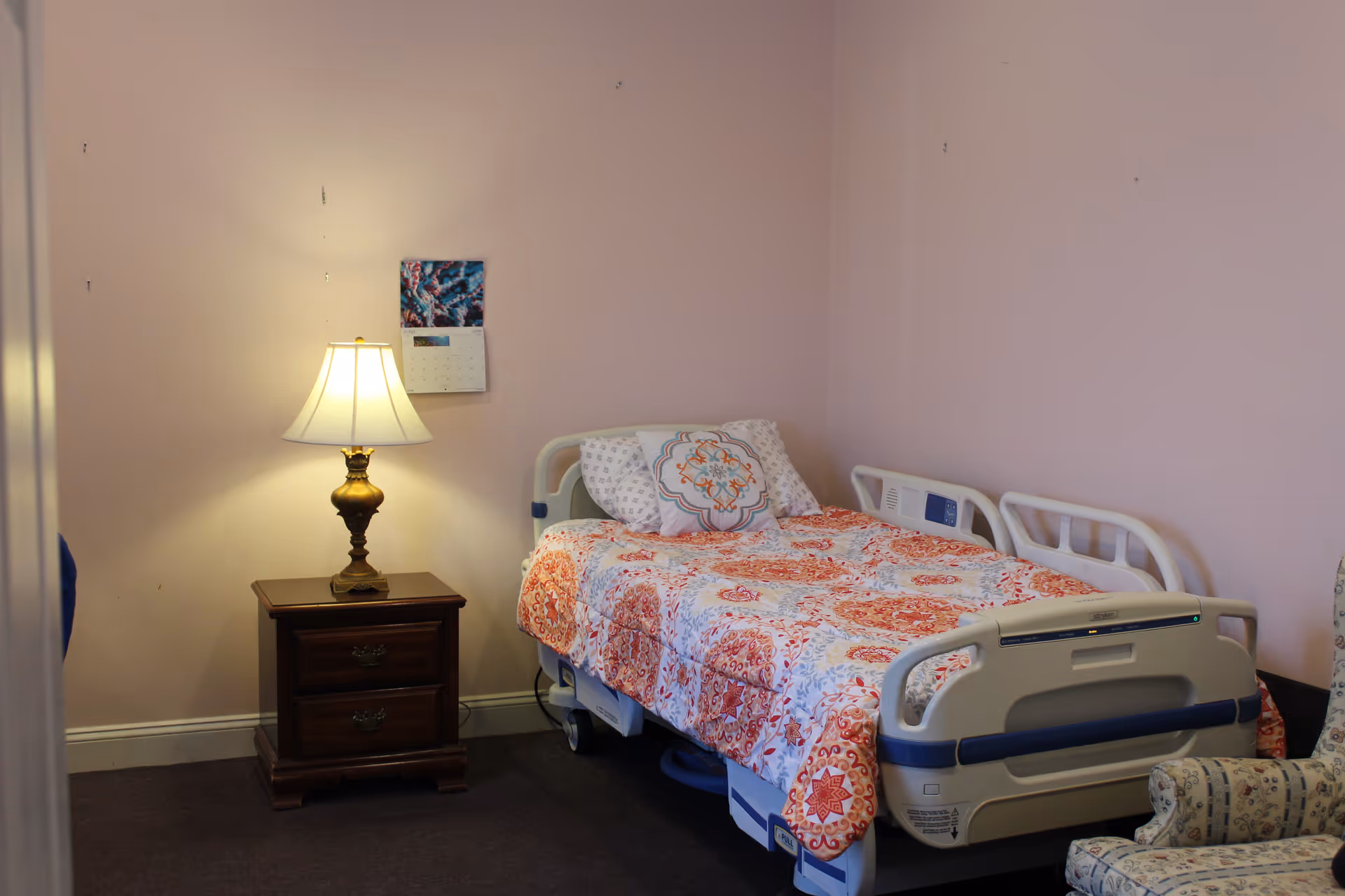 A small bedroom with a hospital-style adjustable bed covered in patterned bedding, a wooden nightstand with a lamp, and an upholstered chair against pink walls.