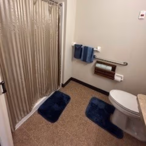 A bathroom with a beige shower curtain, two blue bath mats on the floor, a white toilet with a blue rug in front of it, a towel rack with two blue towels, and a wall-mounted toilet paper holder with extra rolls.