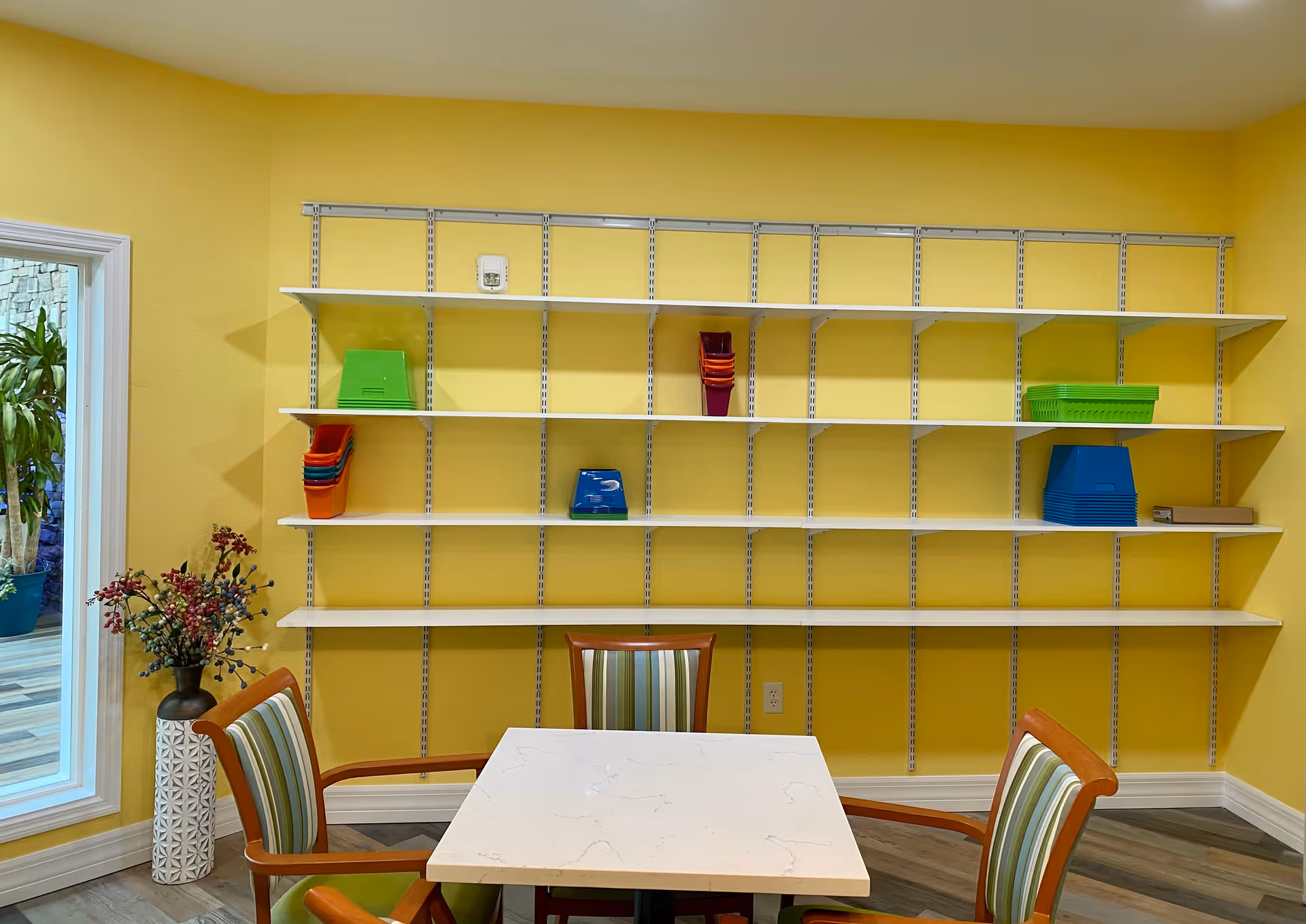 Yellow-walled room with empty white wall-mounted shelves, a table with four striped chairs, and a potted plant by a window.
