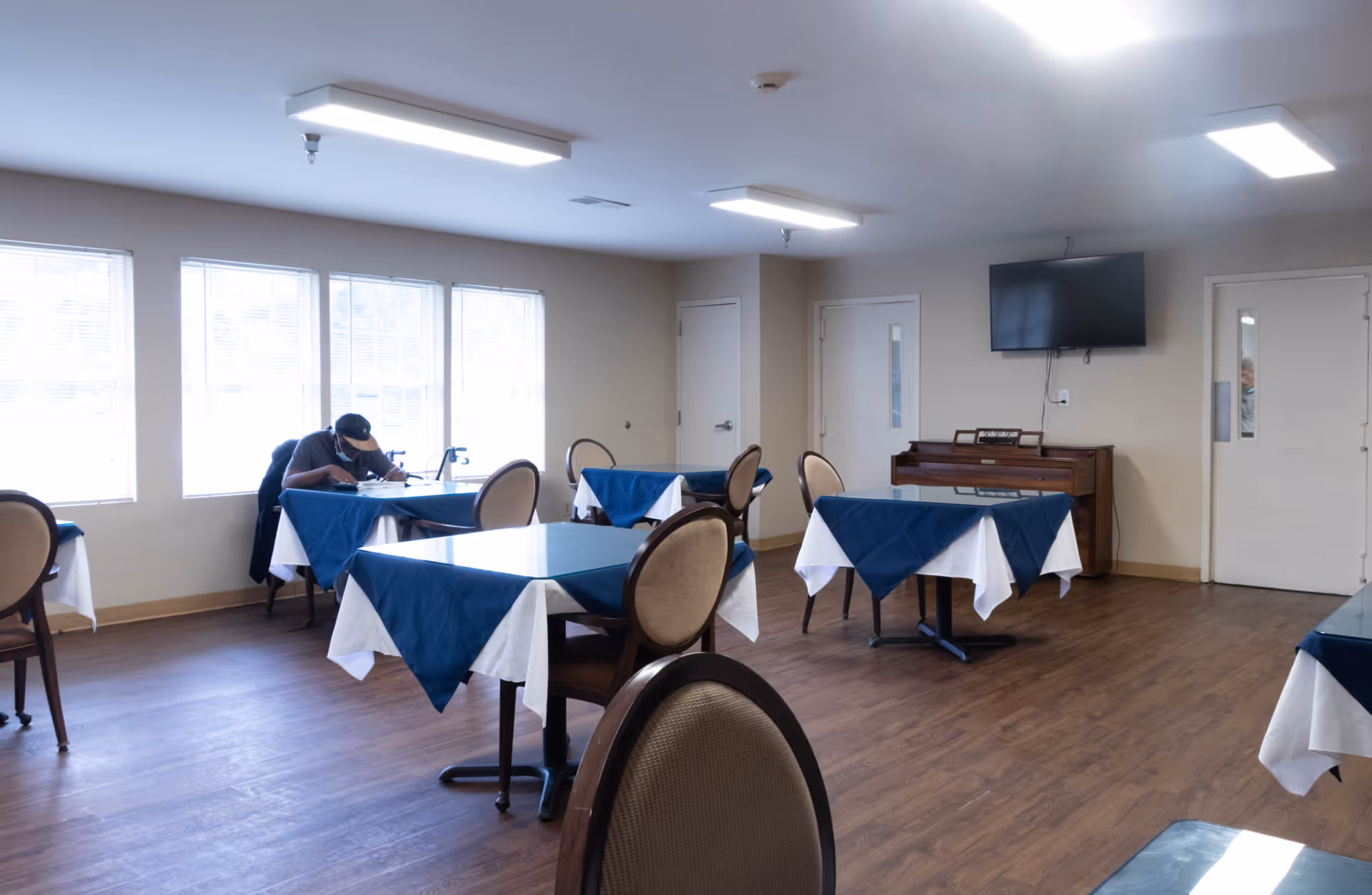 Dining room with tables covered in blue and white tablecloths, chairs, a piano, and a wall-mounted TV.