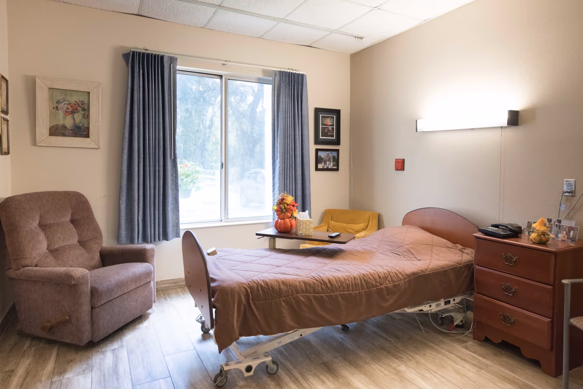 A single hospital-style bed with a brown quilted blanket in a room with wood-look flooring. Next to the bed is a wooden nightstand with a telephone and decorative items. A small table with a tissue box and a fall-themed centerpiece is positioned near a yellow armchair by a window with blue curtains. A brown recliner chair is placed against the opposite wall, which has framed artwork. A bright wall light is mounted above the bed.