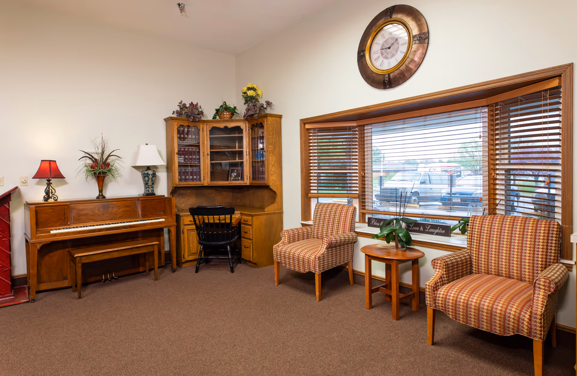 A cozy living room area with a wooden piano on the left, a wooden desk with a black chair in the corner, two patterned armchairs with a small round wooden table between them, a large window with wooden blinds, and a large round wall clock above the window. There are decorative plants and lamps on the piano and desk.