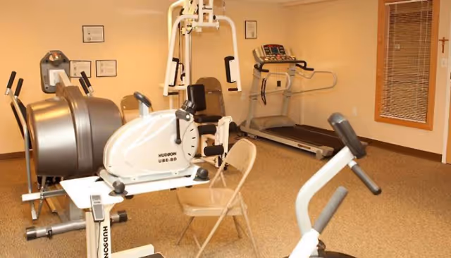 A small fitness room with exercise equipment including a treadmill, a stationary bike, a multi-function weight machine, and a folding chair. The room has beige walls, a window with wooden blinds, and framed certificates or documents on the wall.