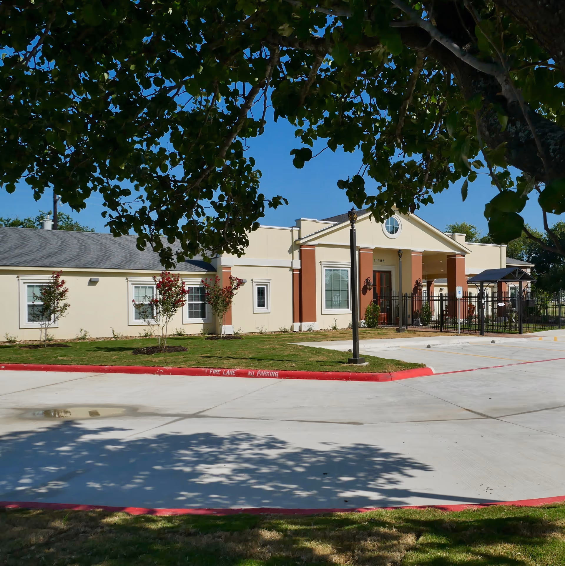 Exterior view of Carol Ann's Home, a single-story building with cream and reddish-brown walls, several windows, and a covered entrance. The foreground shows a paved driveway with red curbs marked 'FIRE LANE NO PARKING' and some green grass and trees casting shadows.