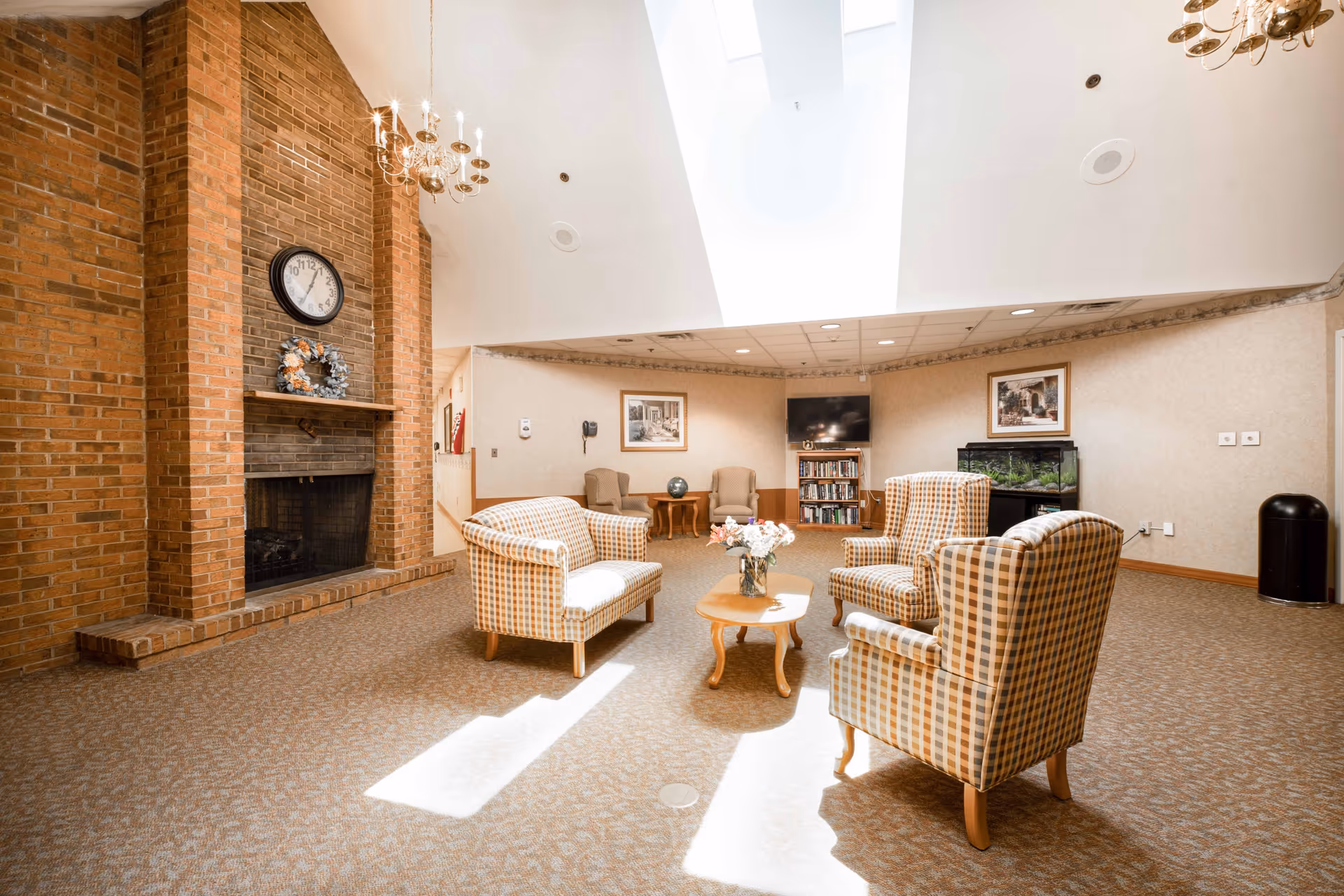 A bright and spacious common area with a high ceiling and skylight. The room features a brick fireplace with a clock and wreath above it, a set of four plaid upholstered chairs arranged around a wooden coffee table with a flower vase, two armchairs with a small table in the background, a television mounted on the wall above a bookshelf, and an aquarium. The walls are decorated with framed pictures and the floor is carpeted.