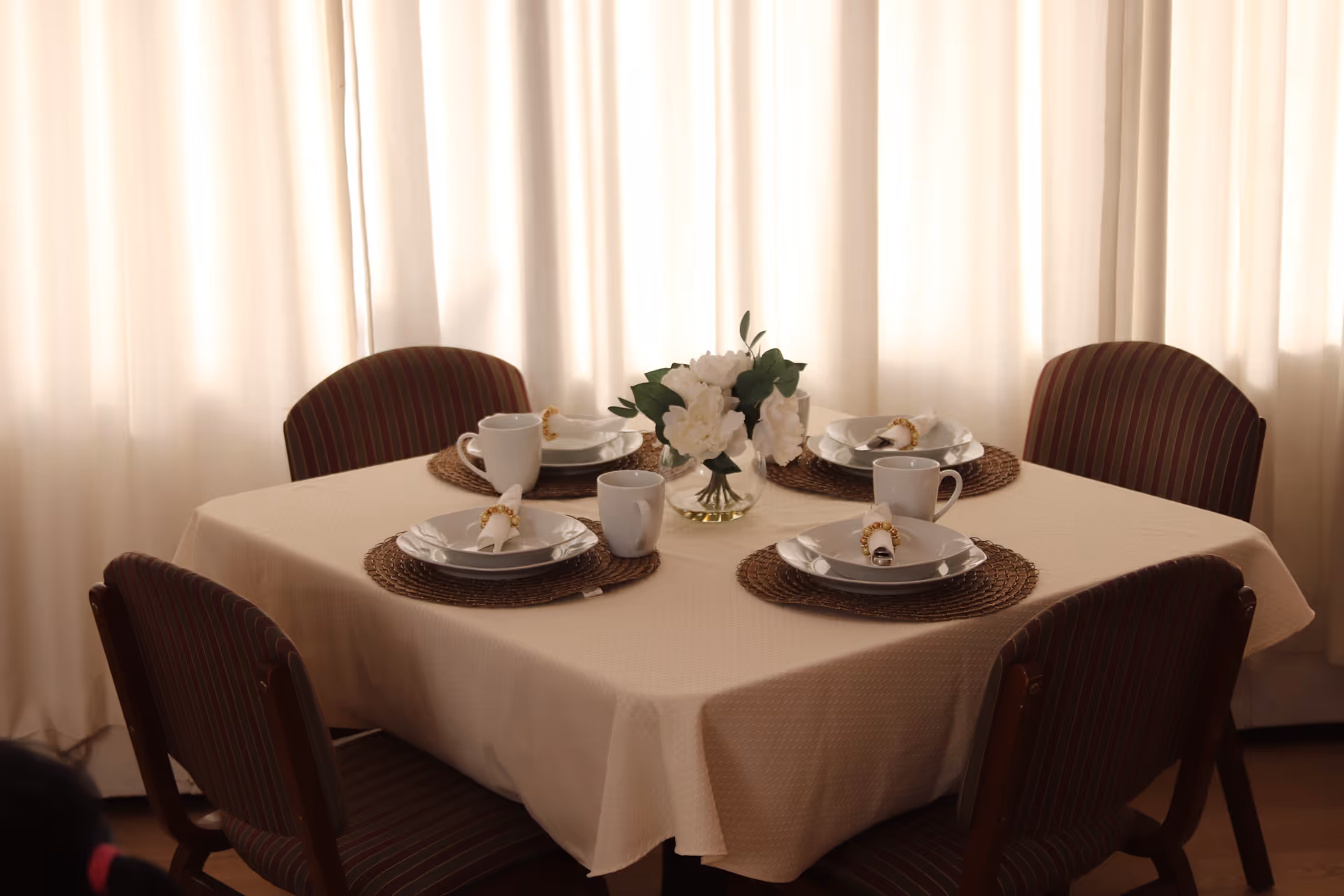 A dining table set for four with plates, mugs, napkins, and a floral centerpiece in front of light curtains.