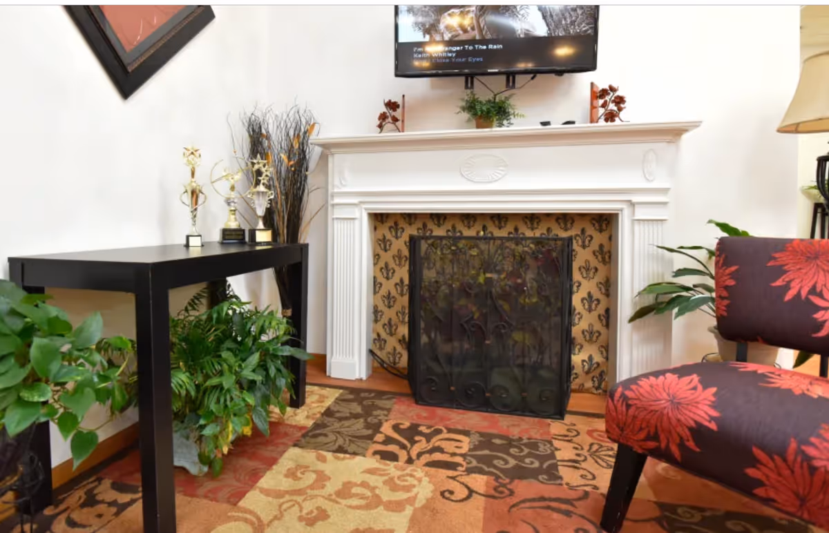 A cozy living room area featuring a white fireplace with a decorative screen in front. On the mantel are small plants and decorative items. Above the fireplace, a flat-screen TV is mounted on the wall. To the left, a black table holds three trophies and a tall vase with decorative branches. Green plants are placed under the table and around the room. On the right, a chair with a dark fabric and large red floral patterns is partially visible. The floor is covered with a multicolored patterned rug.