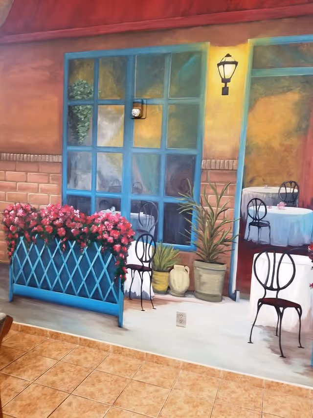 A painted mural on a wall depicting an outdoor café scene with blue window frames, potted plants, a flower planter with pink flowers, and tables with white tablecloths and black chairs. The floor in front of the mural is tiled.