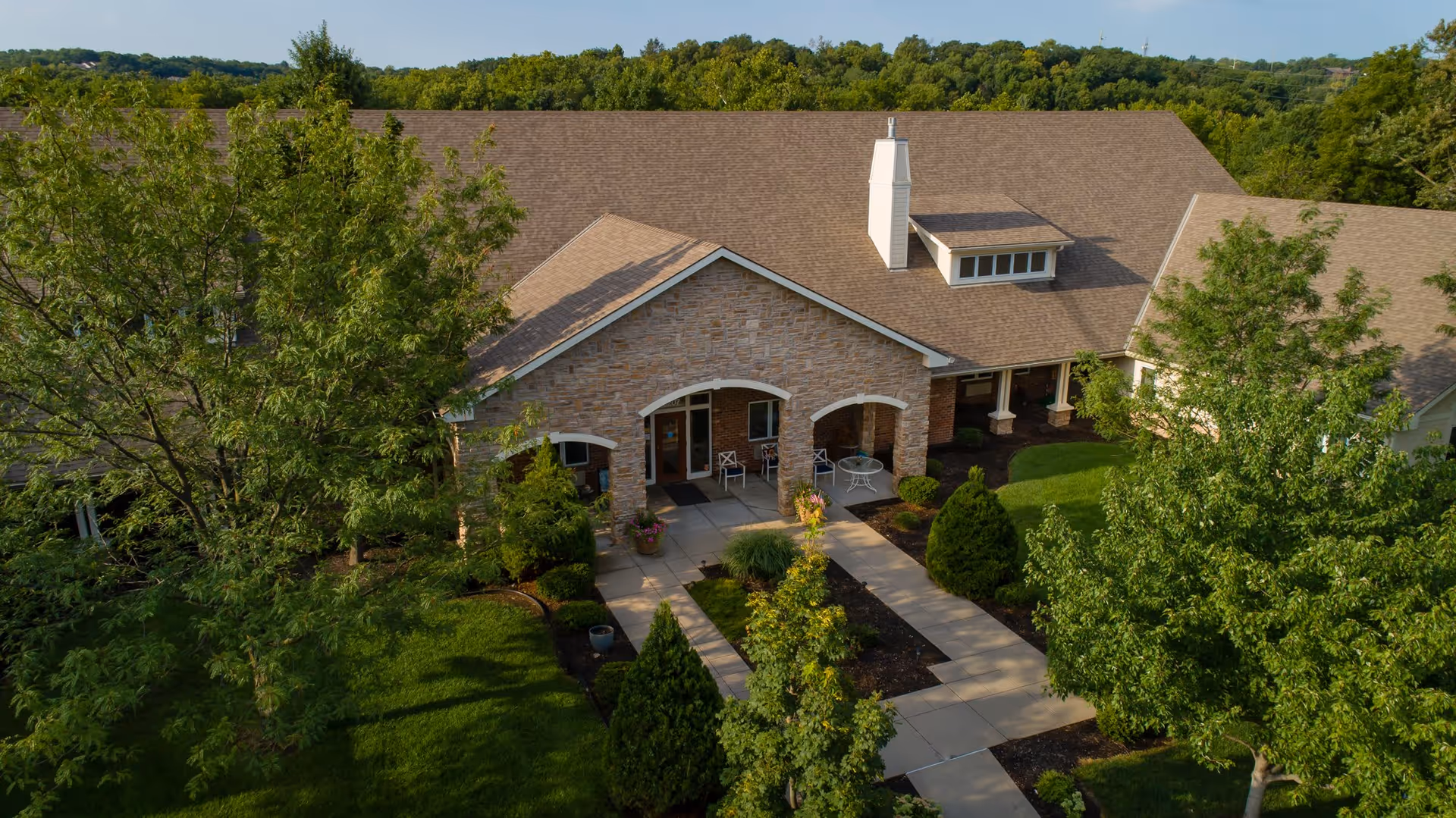 Aerial view of the front entrance of a single-story assisted living facility with a stone facade, a covered porch with chairs and tables, surrounded by well-maintained green lawns, trees, and shrubs.