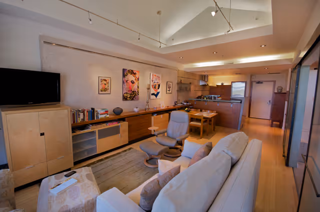 Open-plan living room and kitchen featuring a sofa, recliner, TV, long cabinet with artwork above, and a dining table.