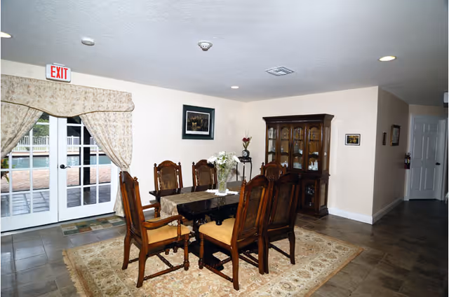 A dining room with a dark wooden dining table and six matching chairs with cushioned seats. The table has a floral centerpiece and a table runner. Behind the table is a wooden china cabinet with glass doors displaying dishes. There is a framed picture on the wall and a small decorative stand with flowers. French doors with floral curtains lead outside to a patio area with a pool visible. The floor is tiled and there is a patterned area rug under the dining table.