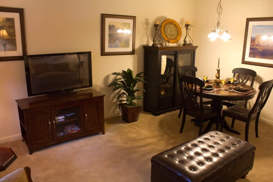 A cozy living and dining area in a senior living facility featuring a flat-screen TV on a wooden stand, a small dining table set for four with chairs, a dark wooden cabinet with decorative items on top, framed artwork on the walls, a potted plant, and a tufted leather ottoman on a carpeted floor.