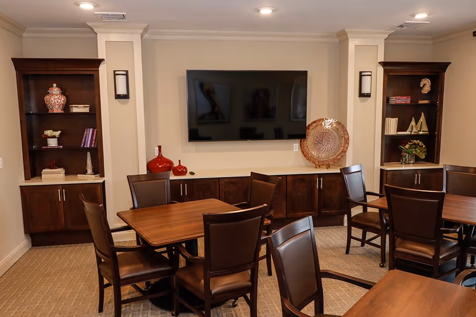 A cozy interior room with multiple wooden tables and chairs arranged for seating. The walls are beige, and there are two built-in dark wood shelves on either side of a mounted flat-screen TV. Decorative items such as vases, books, and a large decorative plate are displayed on the shelves and cabinets below the TV. The room is well-lit with recessed ceiling lights.