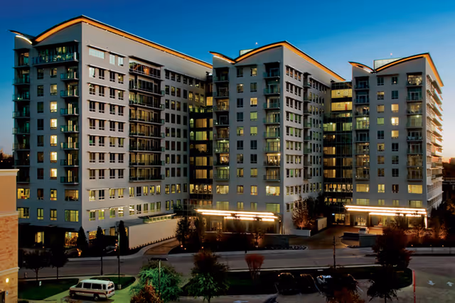 Exterior view of a modern multi-story senior living facility building at dusk with lights on inside and landscaped surroundings including trees and parked cars.