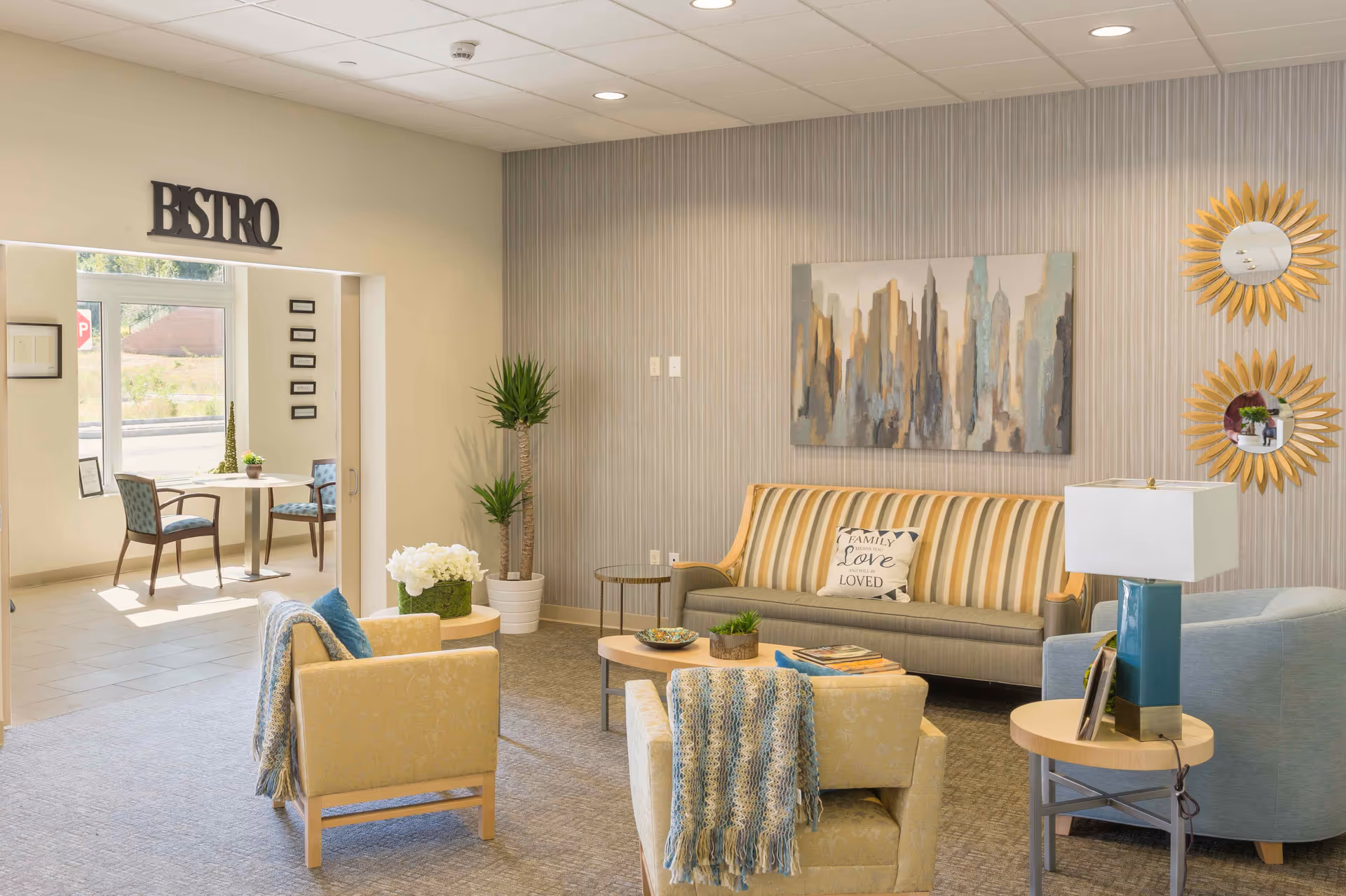 A cozy and well-lit living room area in a senior living facility with a striped sofa, two yellow armchairs with blue and white knitted throws, a blue armchair, and a wooden coffee table with decorative items. The wall has a large abstract cityscape painting and two sunburst mirrors. A side table with a blue lamp and framed photos is also visible. In the background, there is an open doorway leading to a small bistro area with a round table and two chairs near a window.