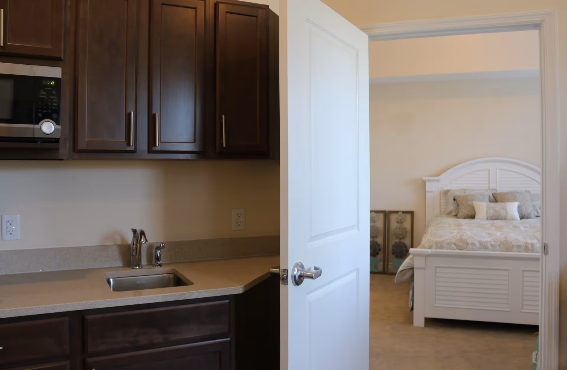 View of a kitchenette area with dark wood cabinets, a microwave, and a sink with a faucet. An open white door leads to a bedroom with a white bed frame and a bed with patterned bedding and pillows. Two framed pictures are leaning against the wall in the bedroom.