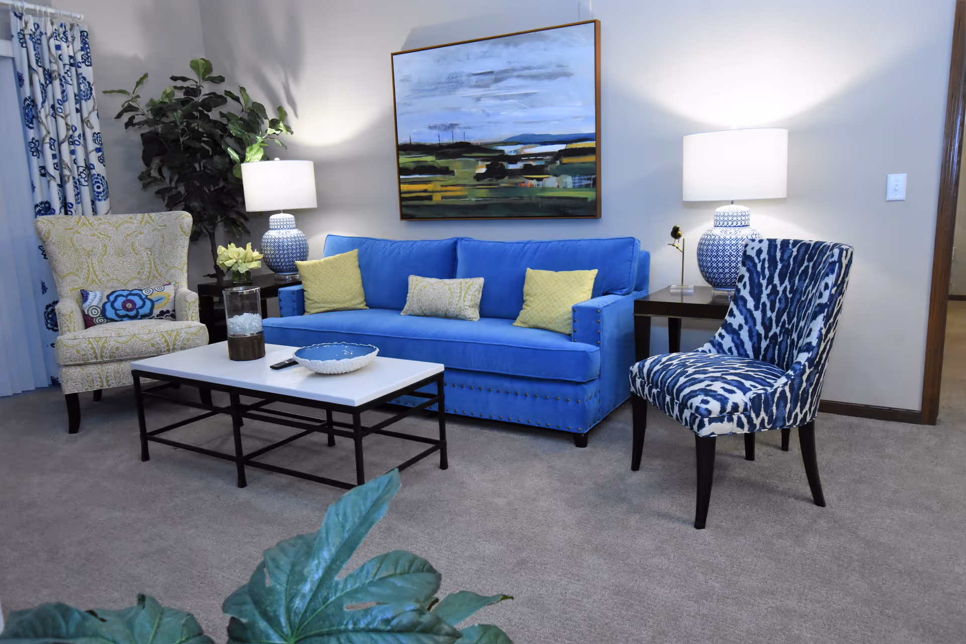 A bright living room with a blue sofa, two patterned armchairs, a coffee table, lamps, and a landscape painting on the wall.