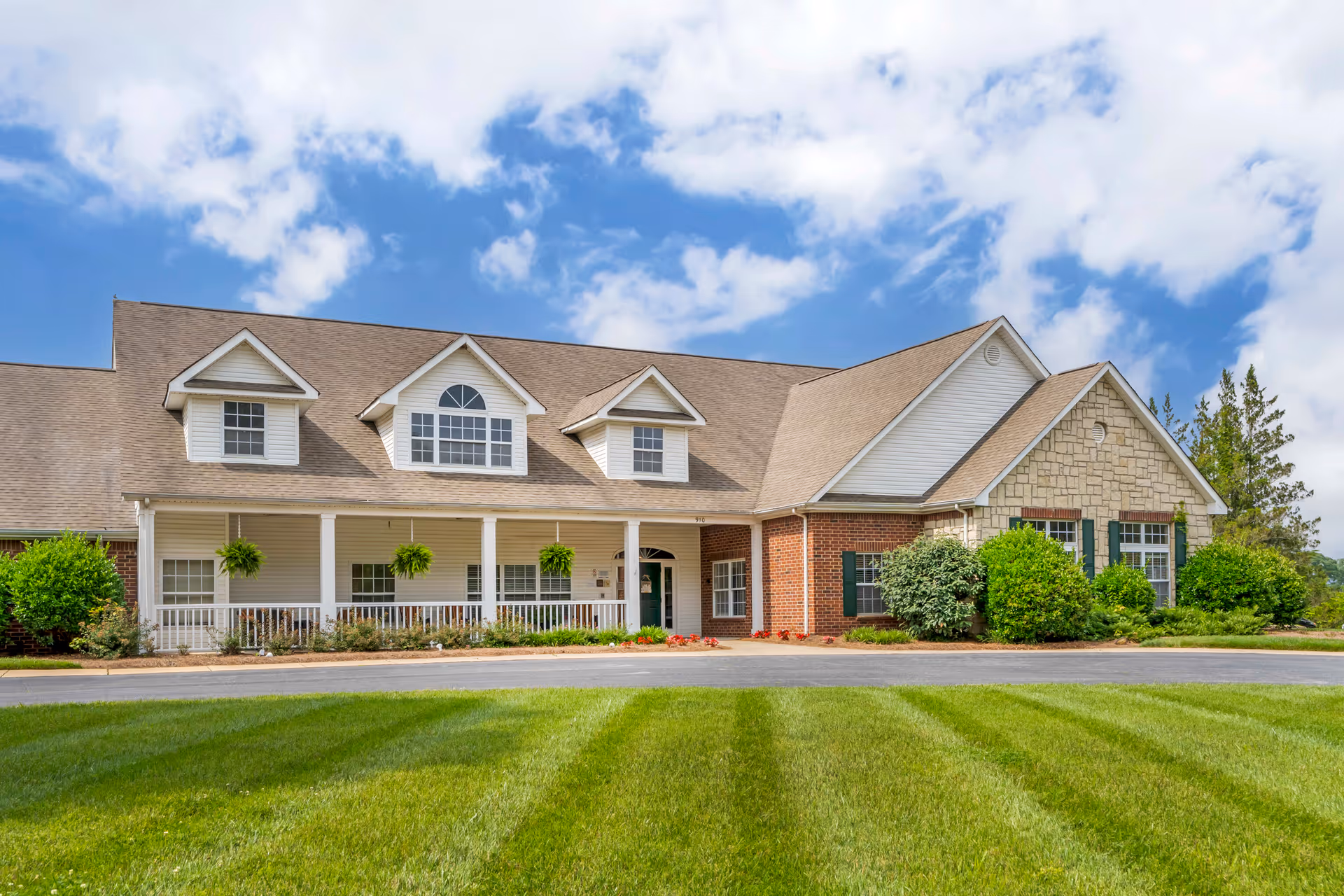 Front exterior view of a single-story building with a combination of brick, stone, and siding. The building has a large porch with white railings and hanging plants, multiple windows, and a well-maintained lawn with green grass under a partly cloudy blue sky.