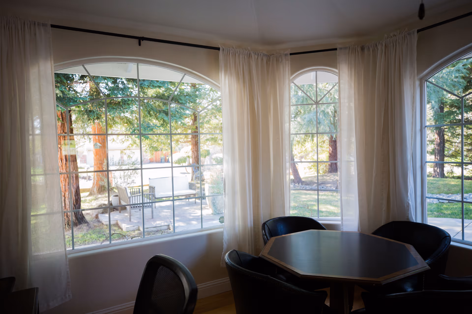 Interior sitting area with an octagonal table and chairs next to large arched windows overlooking trees and a patio.