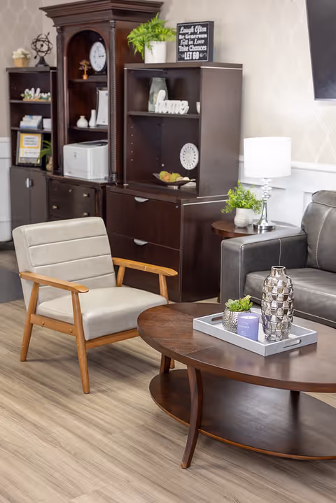 A cozy living room area with a light gray cushioned wooden armchair, a dark brown oval coffee table with decorative items including a silver vase and small plants, a dark gray leather sofa, and dark wood shelving units with decorative items and a white table lamp on a side table.