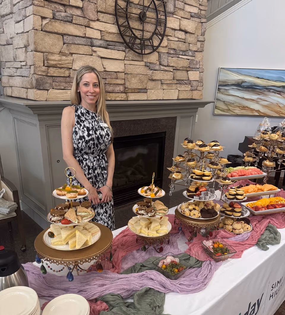 A woman stands behind a buffet table with tiered trays of sandwiches, pastries and fruit set in front of a stone fireplace.