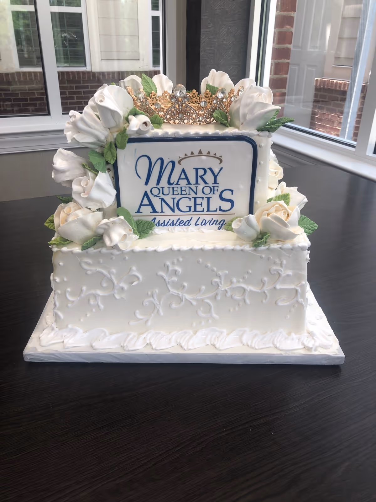 A white decorative cake topped with a gold crown and a sign reading 'Mary Queen of Angels Assisted Living,' displayed on a dark table by windows.