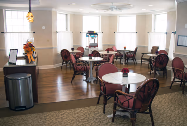 A bright dining area with several round white marble tables surrounded by red cushioned chairs. The room has large windows letting in natural light, wooden flooring in the dining section, and carpeted flooring in the foreground. There is a small counter with a flower arrangement and a menu on the left side, and a popcorn machine near the windows in the background.