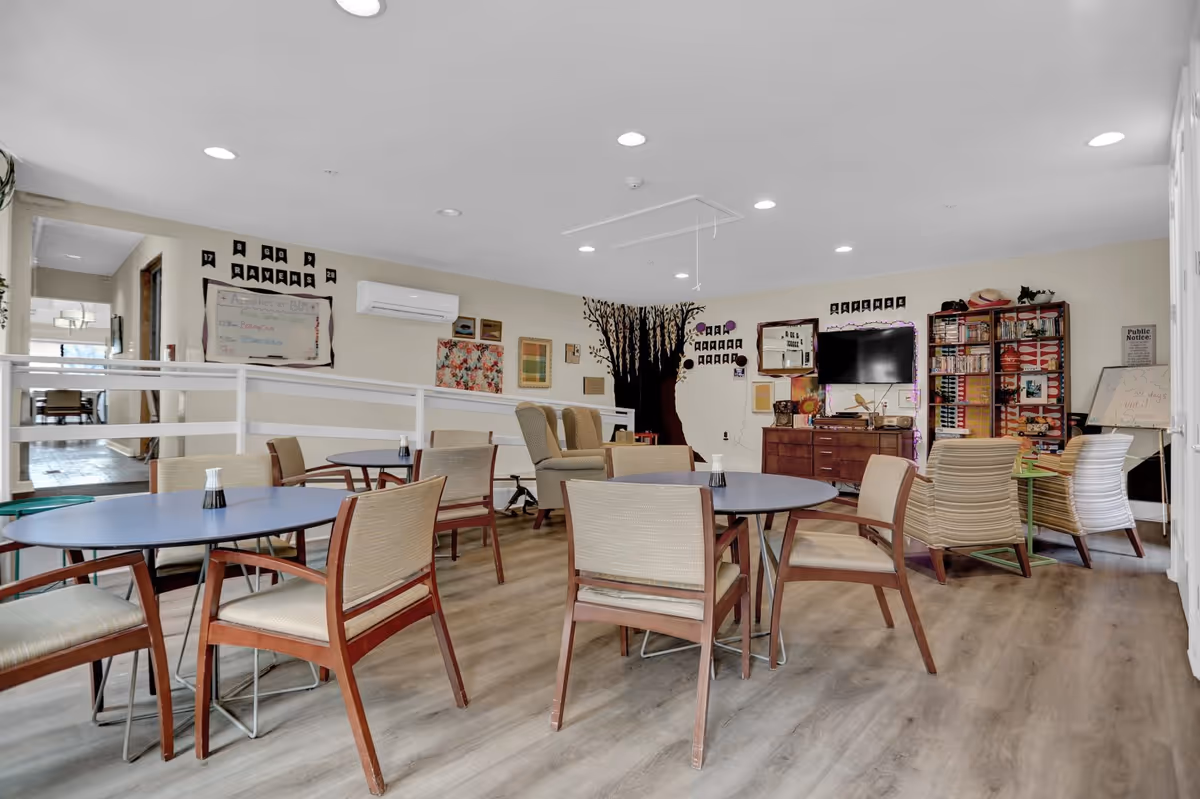 A spacious common area in Broad Creek Manor Assisted Living featuring several round tables with chairs arranged around them. The room has light-colored walls decorated with artwork and a large tree mural. There is a TV mounted on the wall above a wooden cabinet, and a bookshelf filled with books and games. Comfortable armchairs are placed near the TV, and the floor is covered with light wood-style flooring. The room is well-lit with recessed ceiling lights.