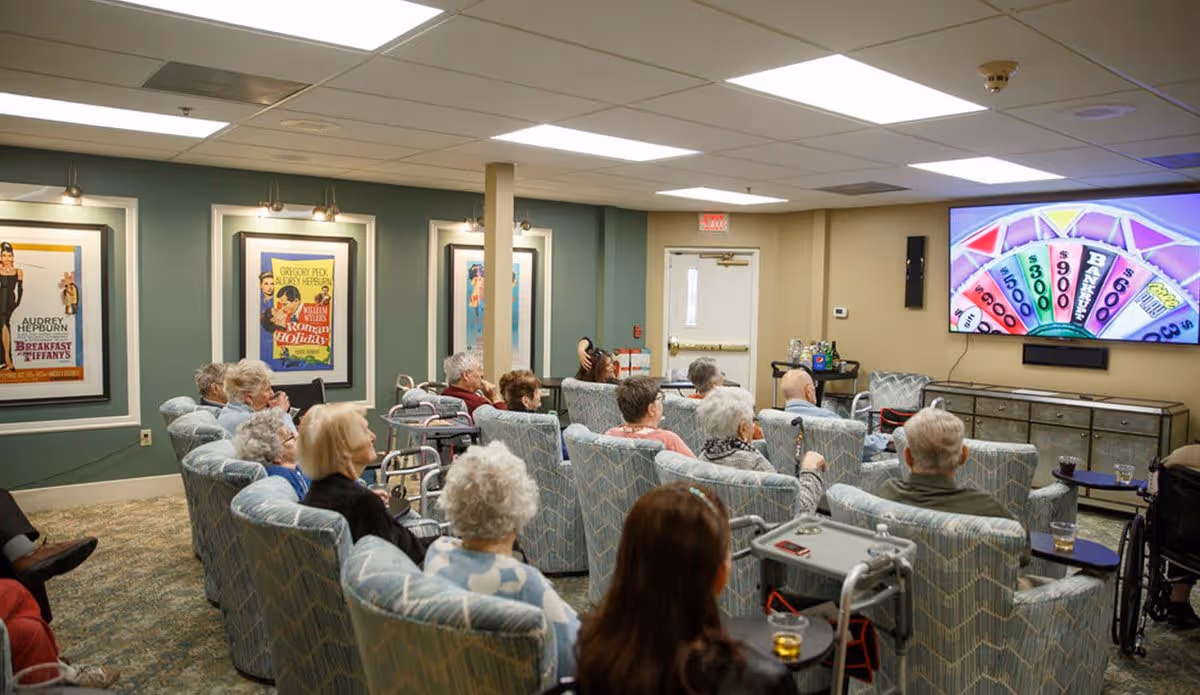 A group of elderly people seated in comfortable armchairs in a common room, watching a large television screen displaying a colorful game show wheel. The room has green walls adorned with framed classic movie posters and a carpeted floor.