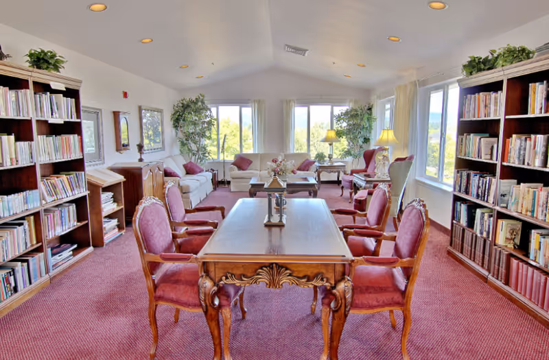 Bright common room with bookshelves, a carved wooden table surrounded by upholstered chairs, sofas, and large windows overlooking greenery.