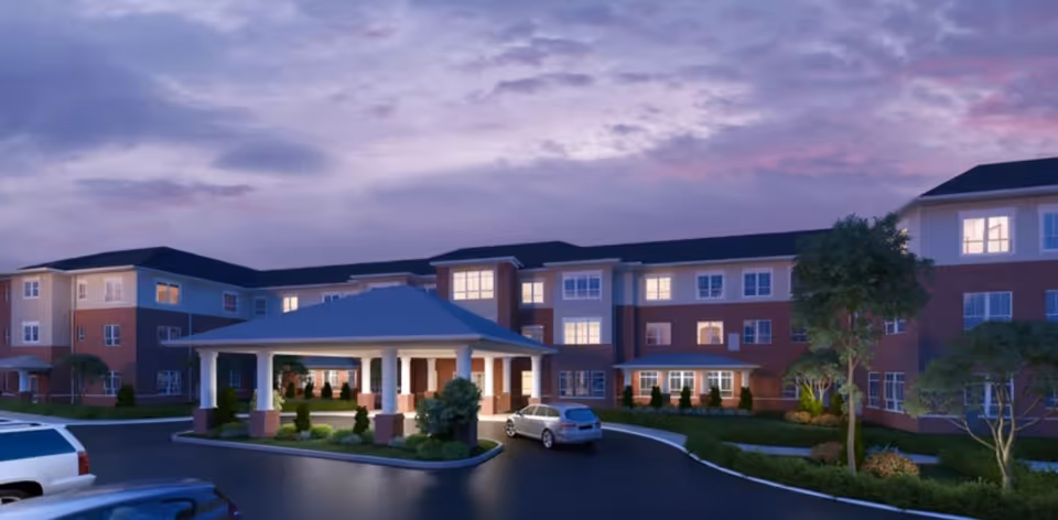 Front exterior of a three-story senior living facility at dusk with a covered porte-cochere and parked cars.