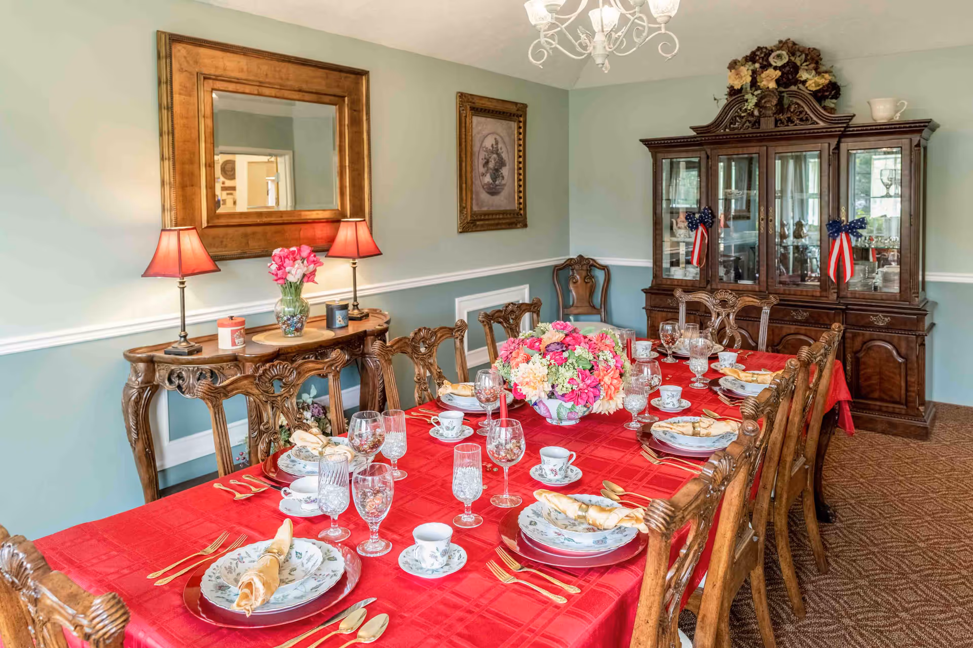 A formal dining room with a long wooden table covered with a red tablecloth, set with floral-patterned plates, cups, crystal glasses, and gold-colored cutlery. A large floral centerpiece decorates the middle of the table. The room features ornate wooden chairs, a wooden sideboard with two lamps and a vase of flowers, a large mirror above the sideboard, a framed picture on the wall, and a wooden china cabinet with glass doors and decorative items inside. The walls are painted light green with white trim, and a chandelier hangs from the ceiling.