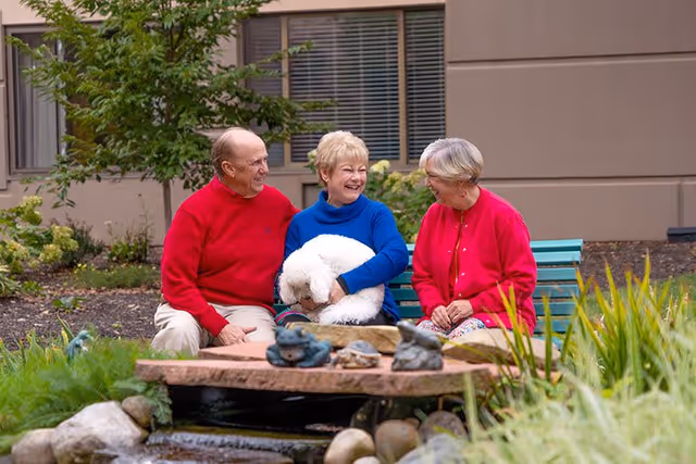 Three elderly people sitting on a bench outdoors near a small pond with decorative rocks and sculptures. One woman in the center is holding a white fluffy dog. They are smiling and engaged in conversation, with greenery and a building in the background.