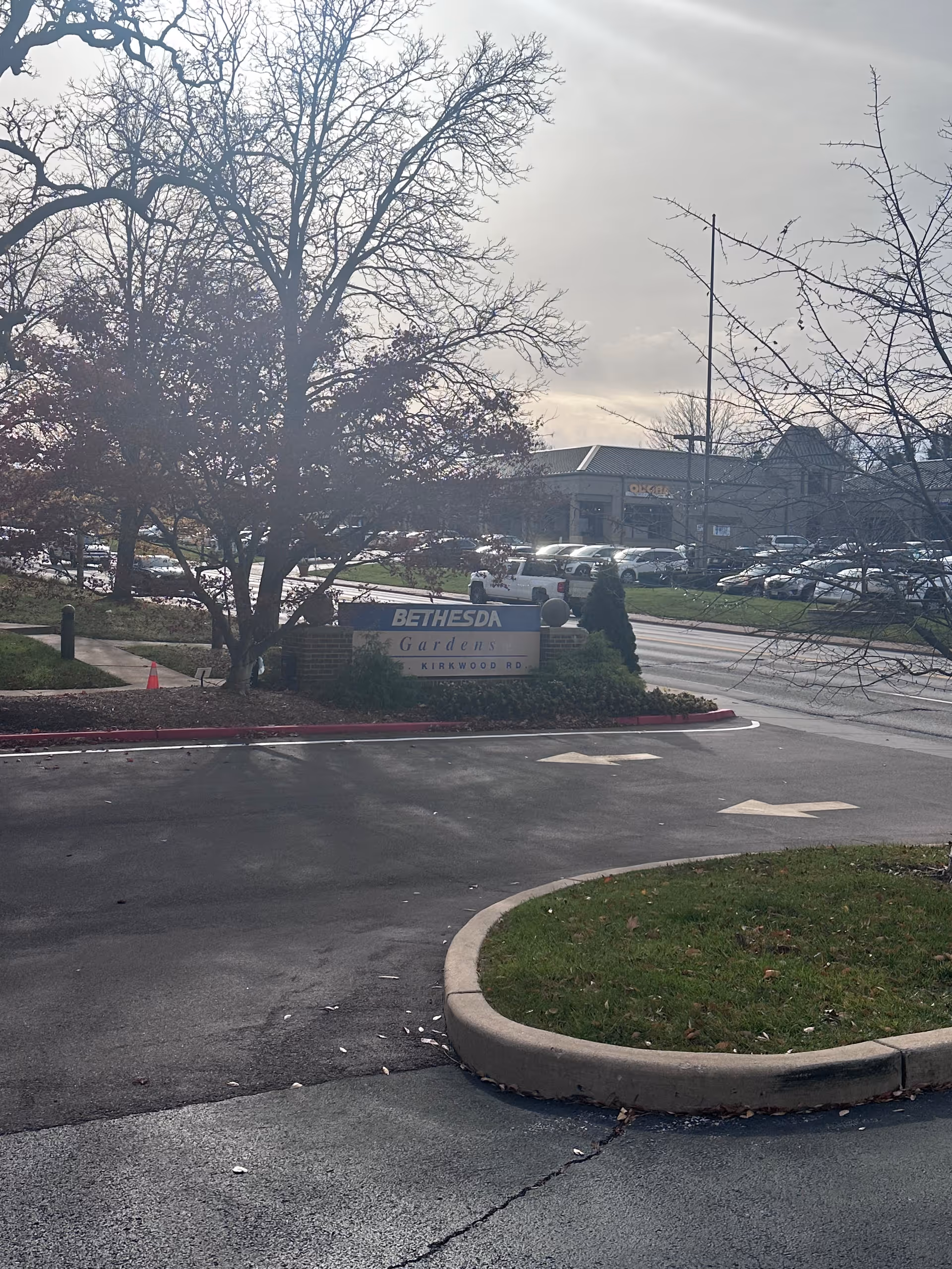 Outdoor view of the entrance area to Bethesda Gardens facility with a sign displaying the name 'Bethesda Gardens Kirkwood Rd'. There are leafless trees, a curved driveway with directional arrows, parked cars in the background, and a partly cloudy sky.