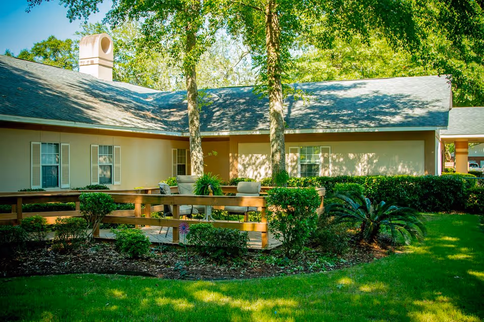 Outdoor patio area at Grandview Retirement Center featuring a wooden deck with cushioned chairs and a table surrounded by green bushes, trees, and a well-maintained lawn in front of a single-story building with beige walls and white window shutters.