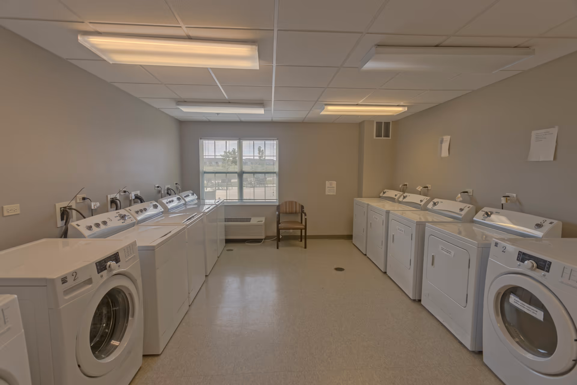 A laundry room with multiple white washing machines and dryers lined up against both side walls. There is a window at the far end of the room with blinds partially open, and a single wooden chair is placed beneath the window. The room has a tiled floor and fluorescent ceiling lights.