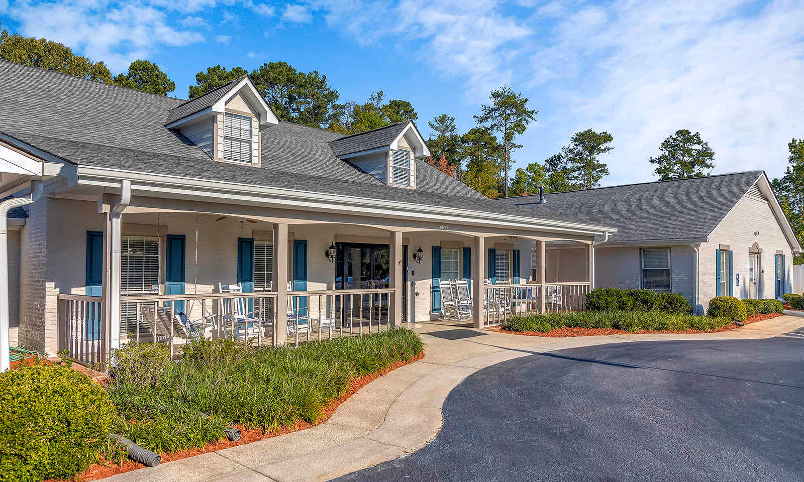 Exterior front view of The Harbor at Douglasville's single-story light-colored building with a covered porch, rocking chairs, blue shutters, landscaping and a curved driveway.