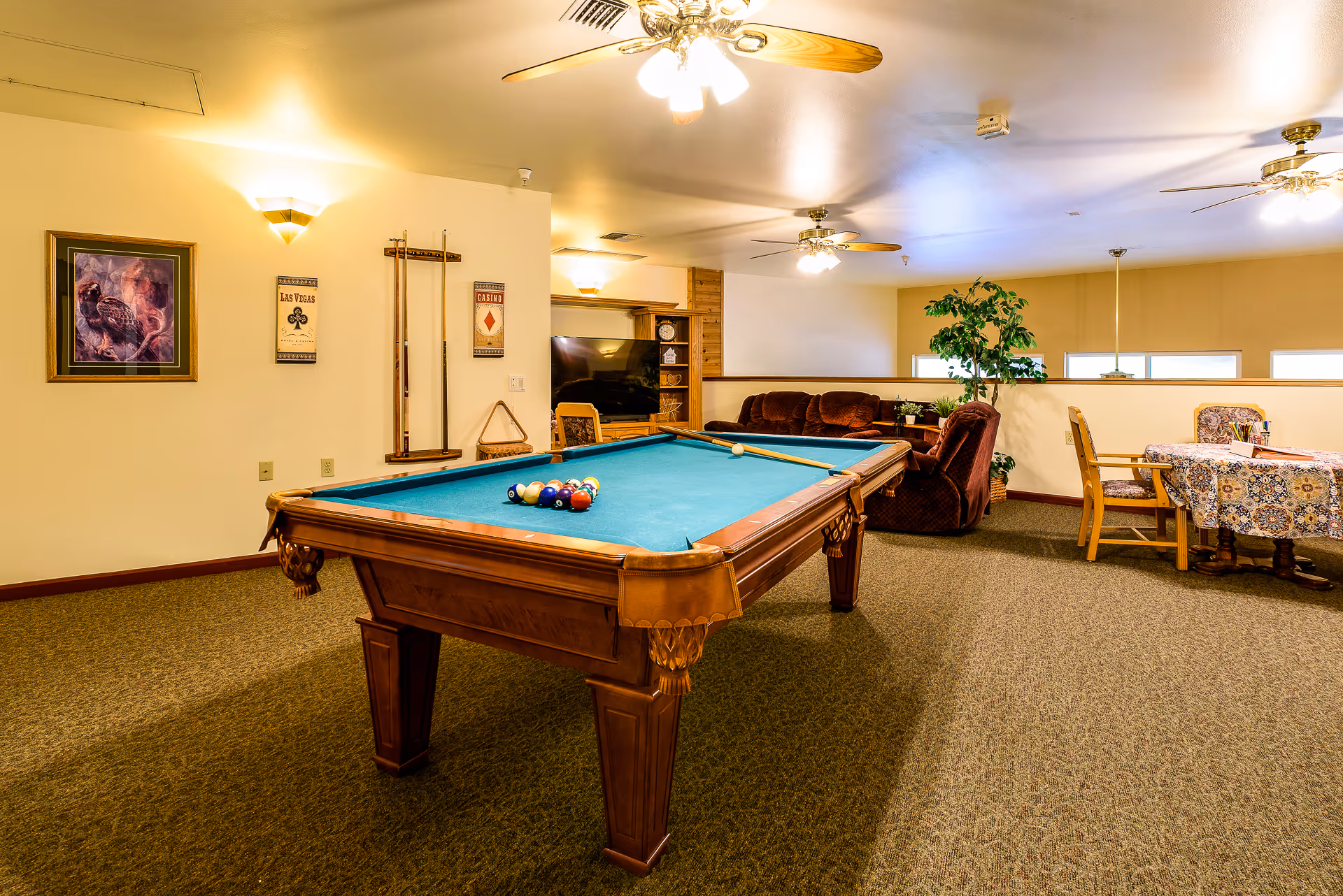 A recreational room featuring a pool table with billiard balls and cues, a seating area with a brown couch and recliner, a TV, a table with chairs covered with a patterned tablecloth, and ceiling fans with lights. The room has beige walls and carpeted flooring.