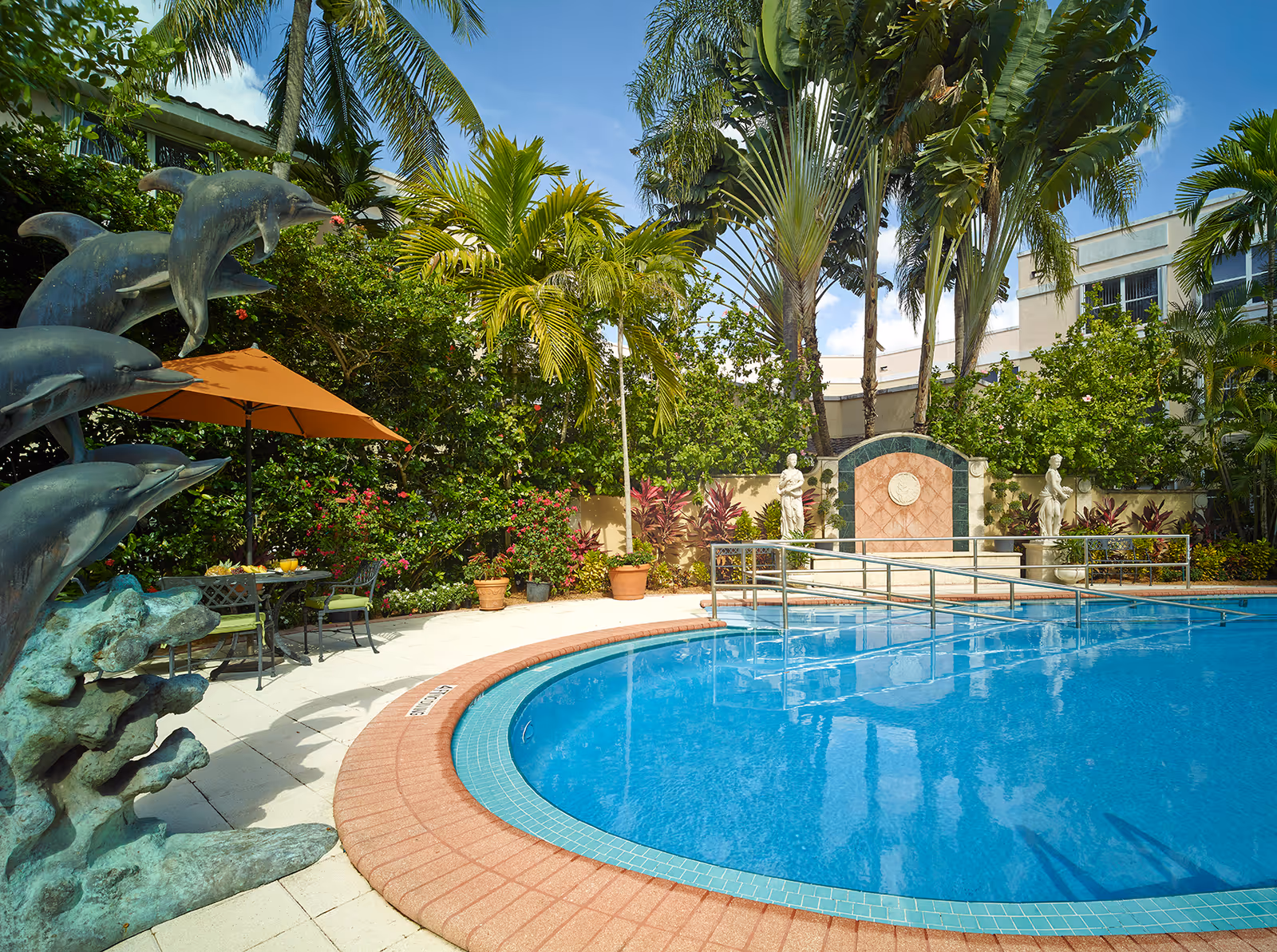 Sunlit outdoor pool area with palm trees, umbrellas, and statues surrounding a curved pool.