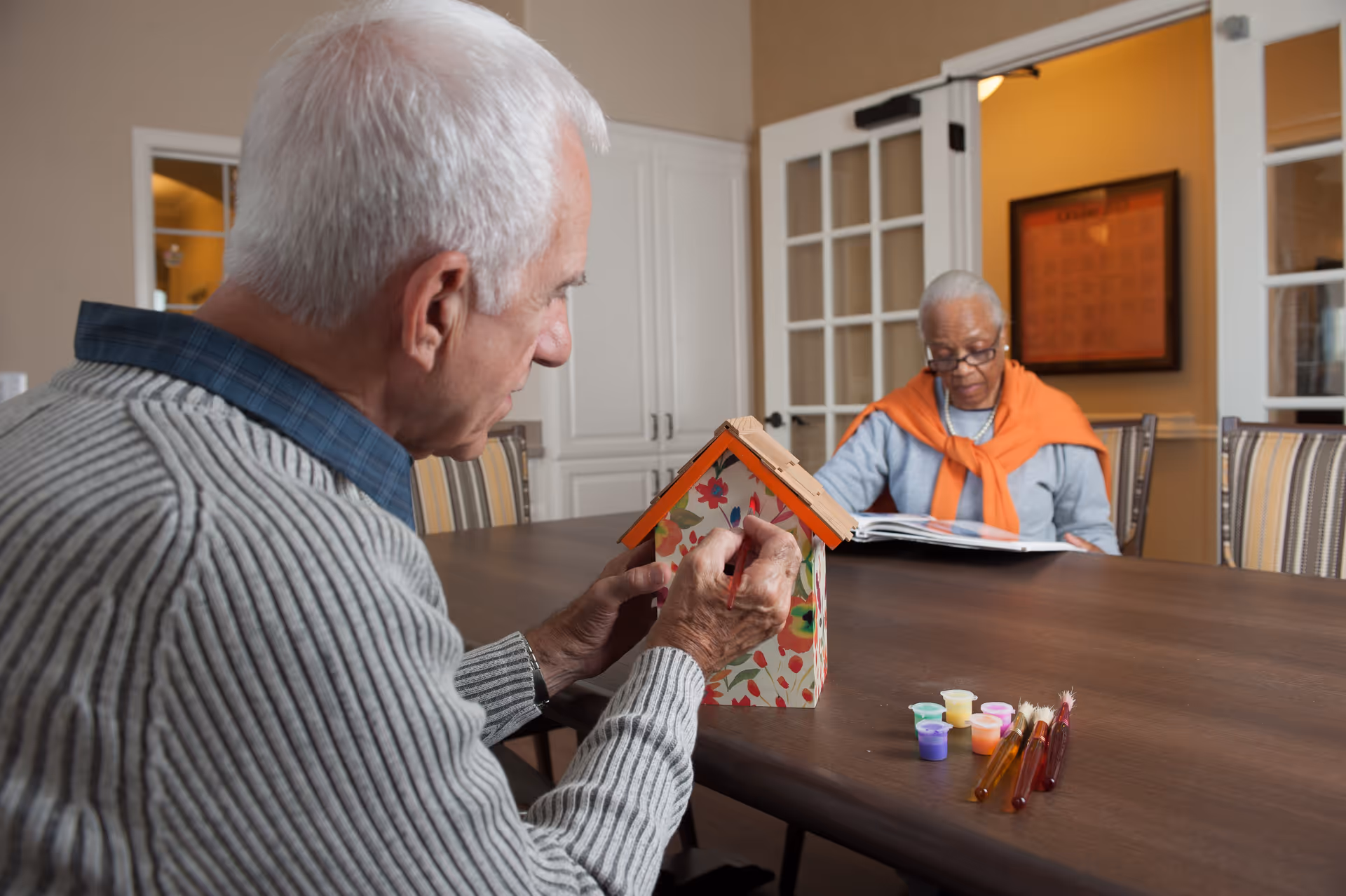 An elderly man painting a decorative birdhouse at a table while an elderly woman sits across from him reading a book in a well-lit room with white cabinets and glass-paneled doors.