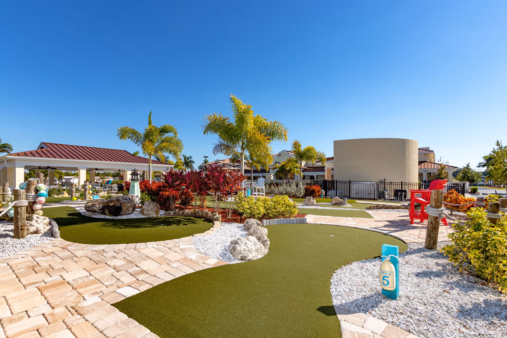 Outdoor mini golf course with artificial turf, palm trees, colorful plants, and a clear blue sky at Naples Estates 55+ Community.