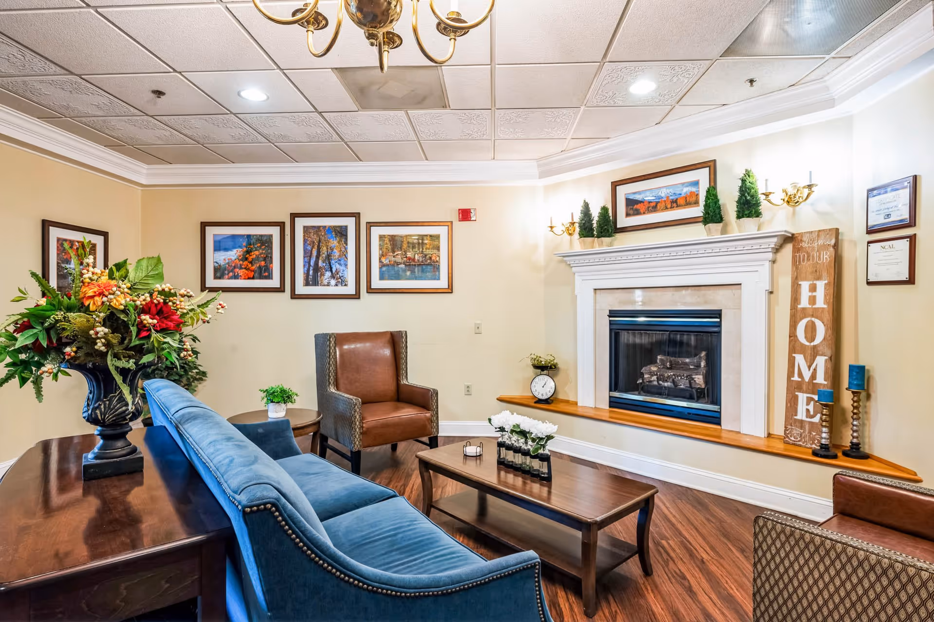 A cozy senior living facility common area with a blue upholstered sofa, a brown leather armchair, a wooden coffee table with small flower vases, and a fireplace with decorative plants and a wooden sign that reads 'HOME'. The walls are adorned with framed landscape photographs and certificates, and there is a chandelier hanging from the ceiling.
