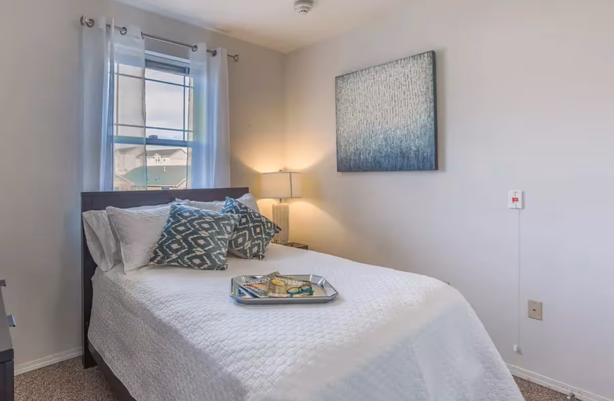 Neat bedroom with a white quilted bed, patterned pillows and a tray, a bedside lamp, window with curtains, and abstract wall art.