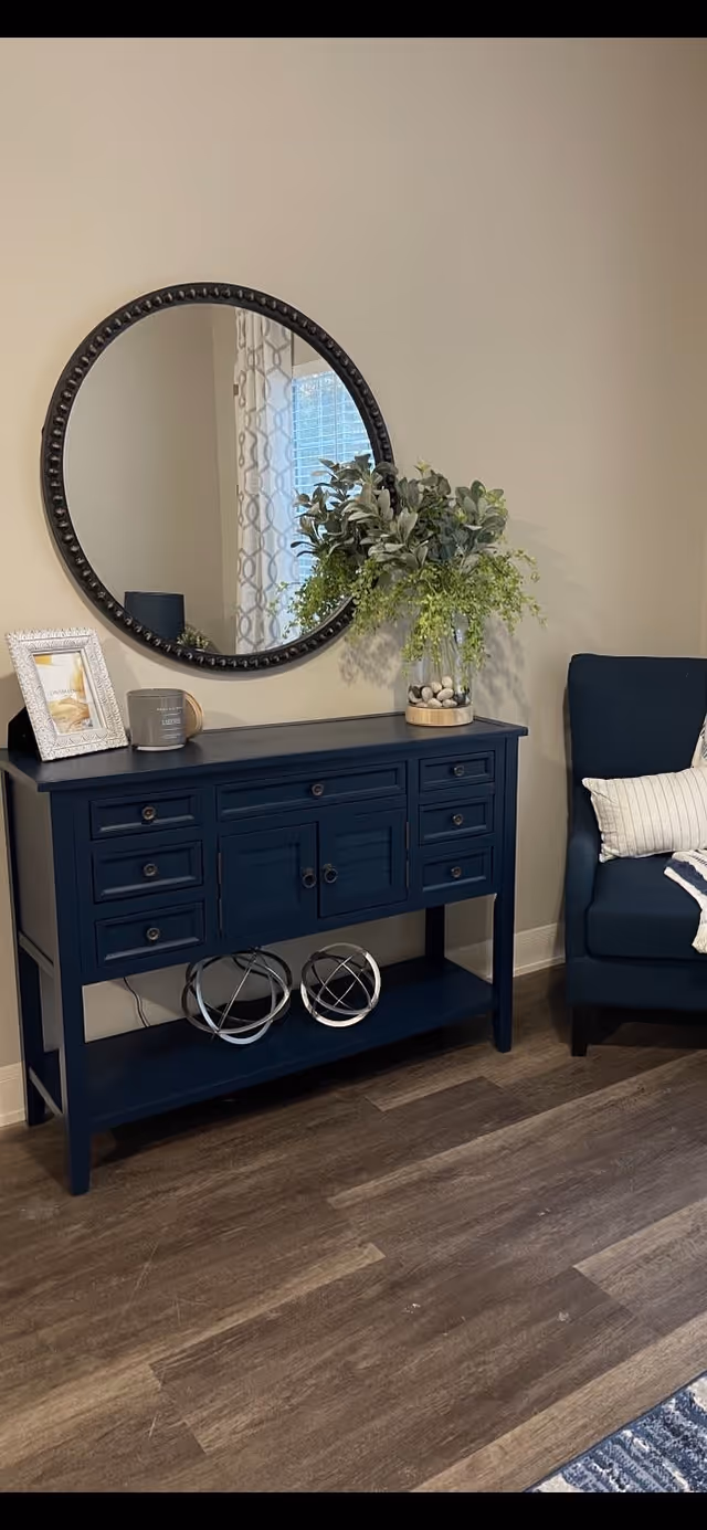 A navy blue console table with multiple drawers and cabinets sits against a beige wall. On top of the table is a round mirror with a dark frame, a framed photo, a candle, and a vase with green leafy plants and decorative stones. To the right of the table is a dark blue upholstered armchair with a white pillow and a striped throw blanket. The floor is wood with a medium brown finish.