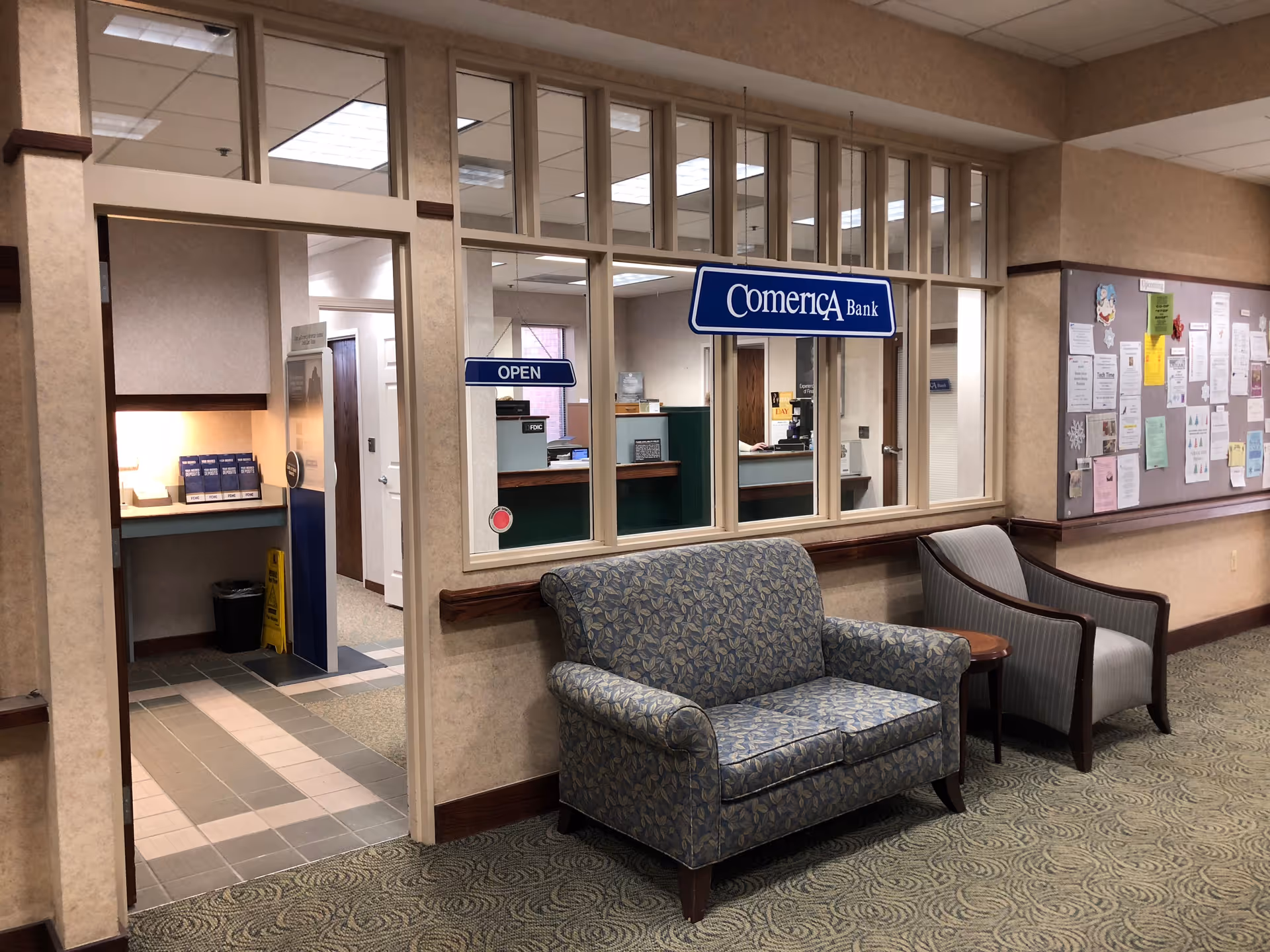 Interior view of a senior living facility hallway featuring a small bank branch with a Comerica Bank sign and an open sign on the window. In front of the bank window, there is a blue patterned loveseat and a gray armchair with a small wooden table between them. To the right, there is a bulletin board with various notices and papers pinned on it. The floor is carpeted with a patterned design, and the walls are beige with wooden trim.
