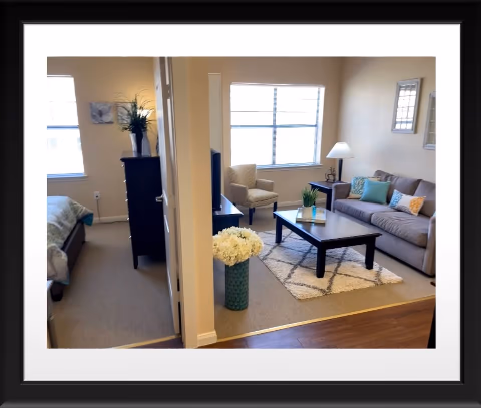 View of a senior living facility apartment showing a living room and a bedroom separated by a partial wall. The living room has a gray sofa with colorful pillows, a coffee table on a patterned rug, an armchair, a side table with a lamp, and a large window. The bedroom has a bed with a green blanket, a tall black dresser with a vase of flowers, and two framed pictures on the wall.