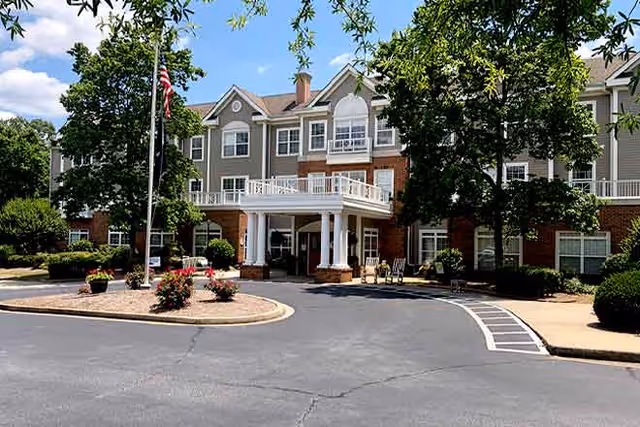 Front exterior view of The Gables senior living facility with a circular driveway, an American flag on a flagpole, and a three-story building featuring a covered entrance supported by white columns. The building is surrounded by trees and landscaping under a partly cloudy sky.