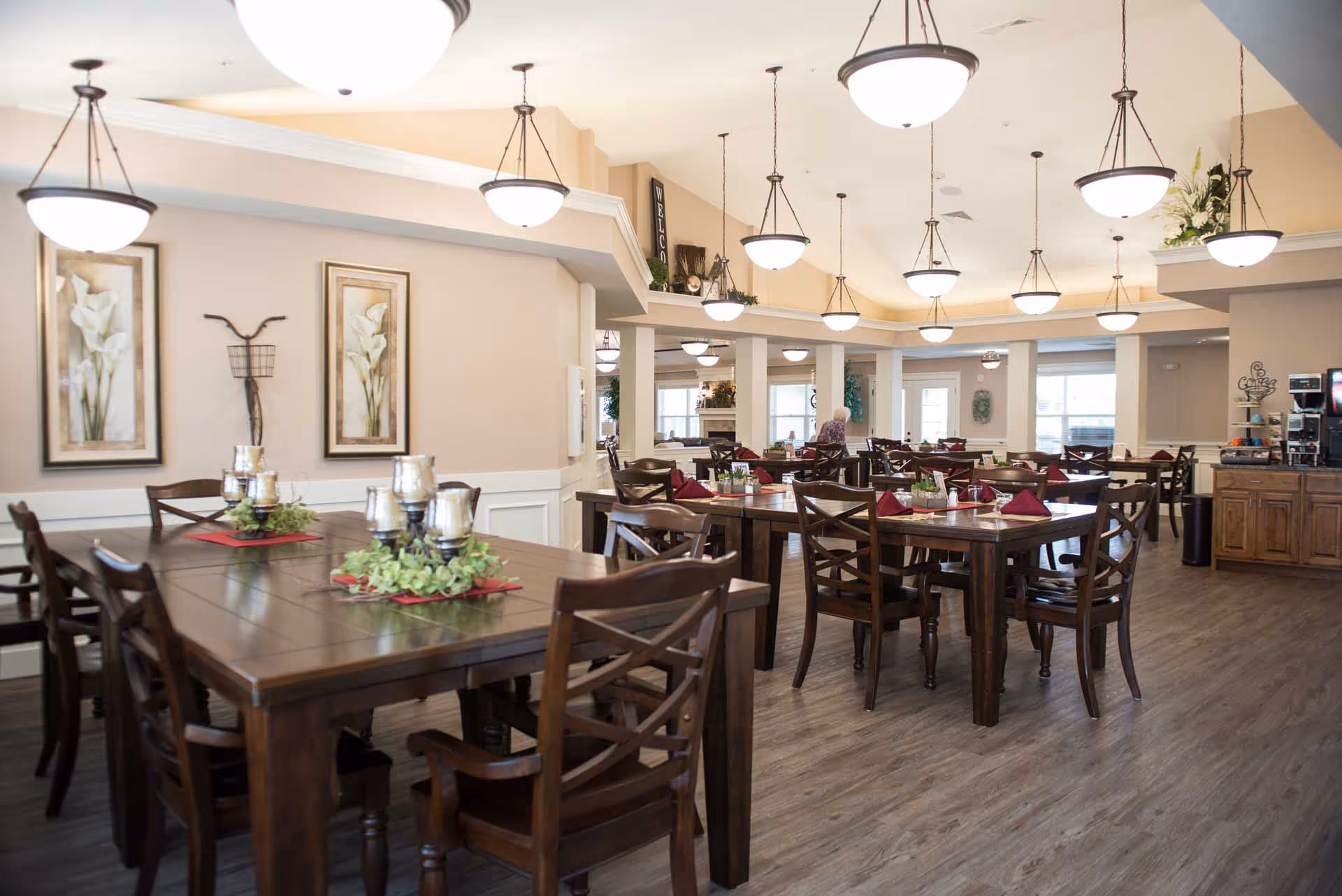 A spacious dining room in an assisted living facility with multiple wooden tables and chairs arranged neatly. Each table is decorated with small plants and candle holders. The room has warm lighting from several hanging ceiling lamps and light-colored walls adorned with framed floral artwork. A coffee and beverage station is visible on the right side, and a person is seen in the background near the windows.