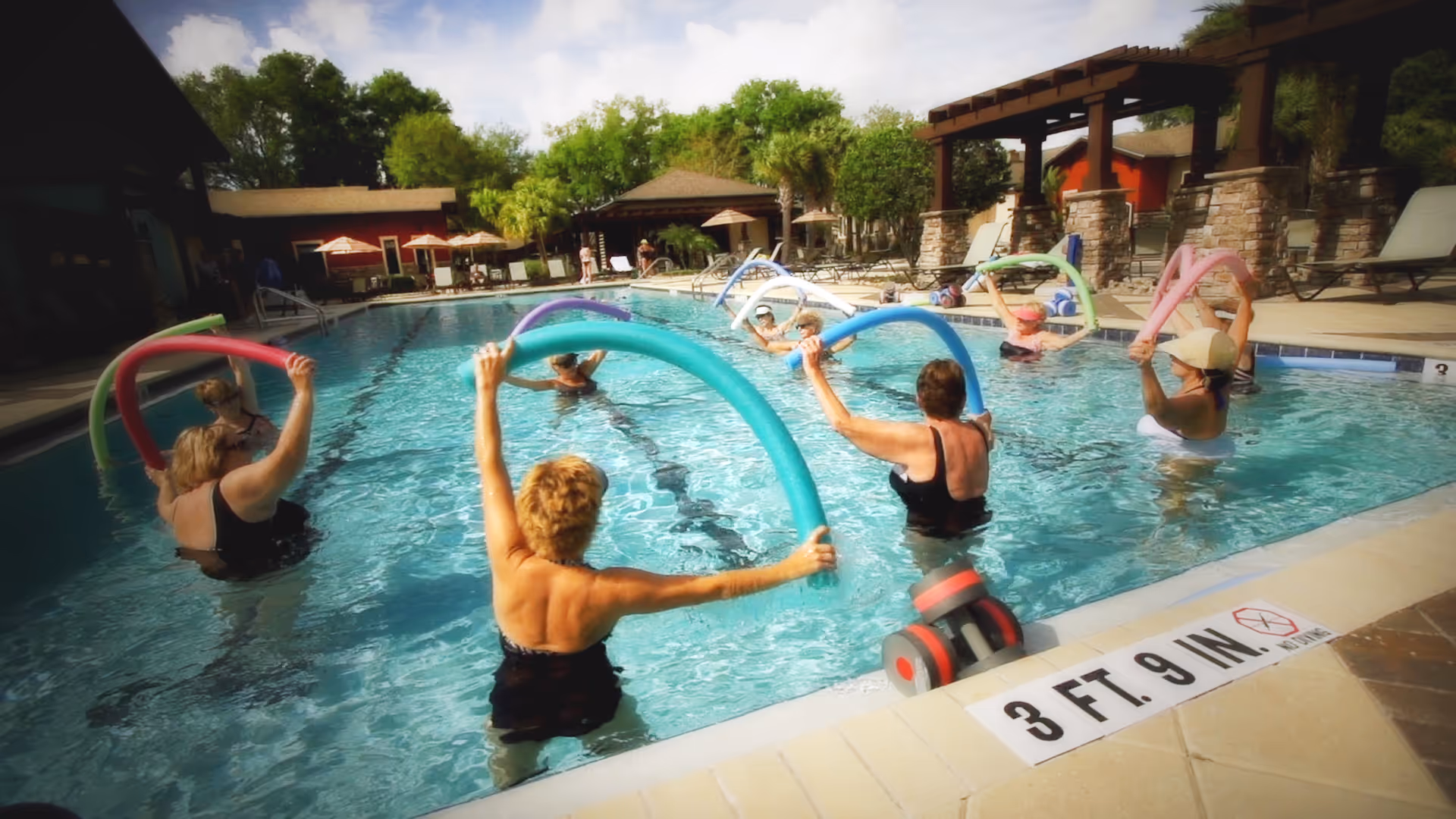 A group of older adults participating in a water exercise class in an outdoor swimming pool, each holding colorful pool noodles above their heads. The pool area is surrounded by lounge chairs, umbrellas, and trees under a partly cloudy sky.