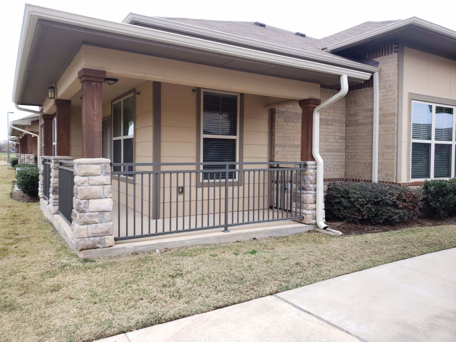Exterior view of a covered porch area with stone and wood pillars, metal railing, and windows on a beige and brick building. There is a concrete walkway and grass lawn in front of the porch.